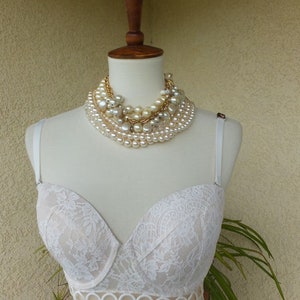 Könnte beinhalten: Weißer Spitzen-BH mit einem gewellten Saum und einem zarten Blumenmuster. Der BH hat einen Push-up-Stil und ist mit einer mehrlagigen Perlenkette verziert.