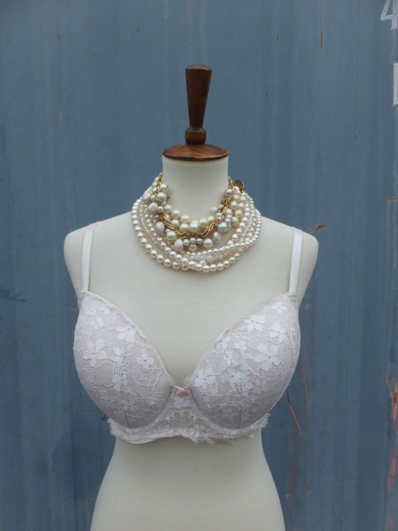 K&ouml;nnte beinhalten: Ein wei&szlig;er Spitzen-BH mit zartem Blumenmuster auf einer wei&szlig;en Schaufensterpuppe. Der BH hat eine rosa Schleife in der Mitte und ist mit mehreren Perlenreihen verziert.