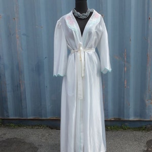 May include: A white long-sleeved robe with a light blue trim and pink floral embroidery on the collar. The robe has a sash tie and is displayed on a mannequin.