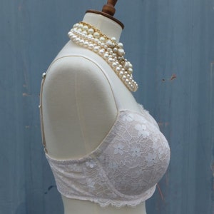 K&ouml;nnte beinhalten: Ein wei&szlig;er Spitzen-BH mit Blumenmuster auf einer wei&szlig;en Schaufensterpuppe. Der BH hat verstellbare Tr&auml;ger und einen Haken- und &Ouml;senverschluss auf der R&uuml;ckseite.