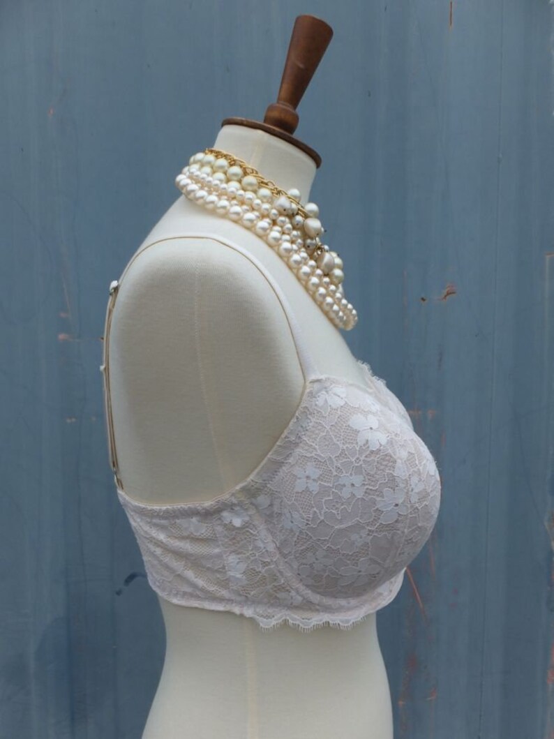 K&ouml;nnte beinhalten: Ein wei&szlig;er Spitzen-BH mit Blumenmuster auf einer wei&szlig;en Schaufensterpuppe. Der BH hat verstellbare Tr&auml;ger und einen Haken- und &Ouml;senverschluss auf der R&uuml;ckseite.