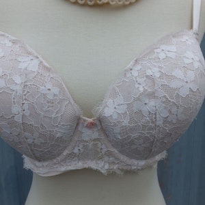 K&ouml;nnte beinhalten: Ein wei&szlig;er Spitzen-BH mit rosa Knopfverschluss. Der BH befindet sich auf einer wei&szlig;en Schaufensterpuppe mit einer Perlenkette.