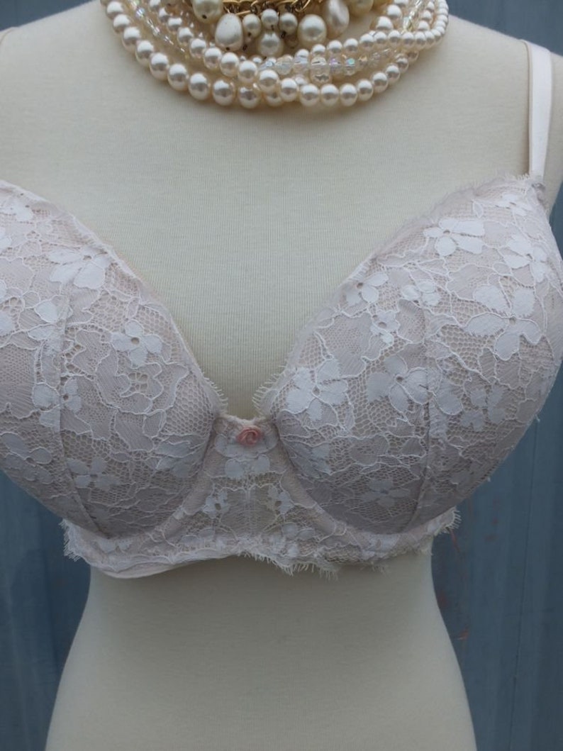 K&ouml;nnte beinhalten: Ein wei&szlig;er Spitzen-BH mit rosa Knopfverschluss. Der BH befindet sich auf einer wei&szlig;en Schaufensterpuppe mit einer Perlenkette.