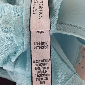 May include: A light blue bra with lace detailing and a pink tag that reads "Victoria's Secret Angels" and includes information about the bra's size, material, and origin. The tag also says "lined demi/demi doubl&eacute;" and "made in India/fabriqu&eacute; en Inde/hecho en India/fabbricato in India/印度制造". The bra size is 34D/D75.