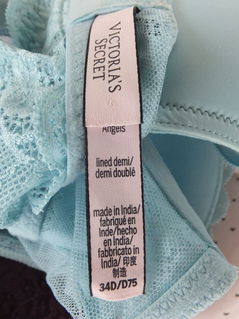 May include: A light blue bra with lace detailing and a pink tag that reads "Victoria's Secret Angels" and includes information about the bra's size, material, and origin. The tag also says "lined demi/demi doubl&eacute;" and "made in India/fabriqu&eacute; en Inde/hecho en India/fabbricato in India/印度制造". The bra size is 34D/D75.