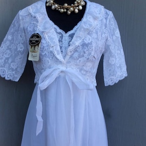 Op de afbeelding: Een witte nachtjapon van kant met een bijpassend kanten jasje. Het jasje heeft een gerimpelde kraag en korte mouwen. De nachtjapon heeft een strik in de taille en een doorschijnende rok. De nachtjapon is op een mannequin met een parelketting.