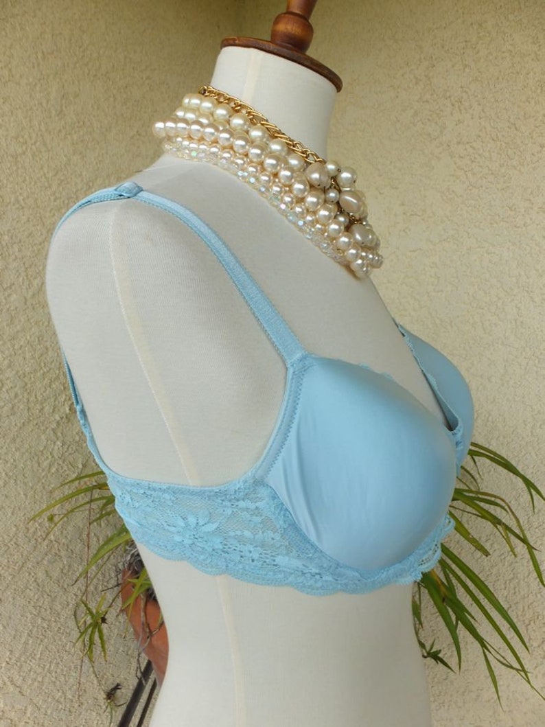 May include: A light blue lace bra with adjustable straps. The bra is on a white mannequin with a pearl necklace.