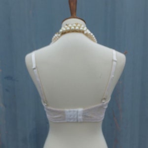 K&ouml;nnte beinhalten: Wei&szlig;er Spitzen-BH mit verstellbaren Tr&auml;gern und Haken- und &Ouml;senverschluss im R&uuml;cken. Der BH befindet sich auf einer wei&szlig;en Schaufensterpuppe mit einer Perlenkette.