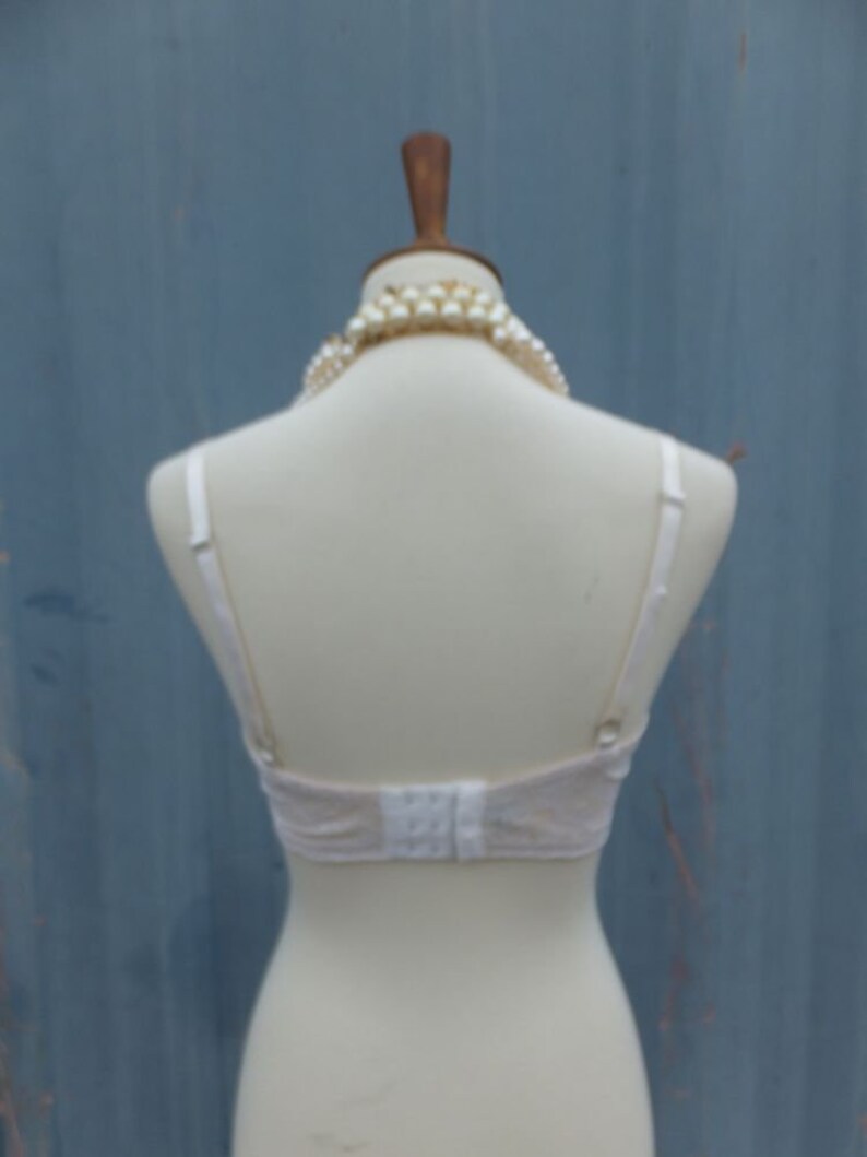 K&ouml;nnte beinhalten: Wei&szlig;er Spitzen-BH mit verstellbaren Tr&auml;gern und Haken- und &Ouml;senverschluss im R&uuml;cken. Der BH befindet sich auf einer wei&szlig;en Schaufensterpuppe mit einer Perlenkette.