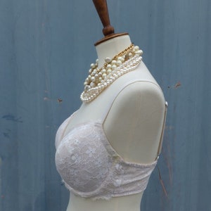 K&ouml;nnte beinhalten: Ein wei&szlig;er Spitzen-BH auf einer wei&szlig;en Schaufensterpuppe mit einer Perlen- und Goldkette. Die Puppe steht vor einem blauen Hintergrund.