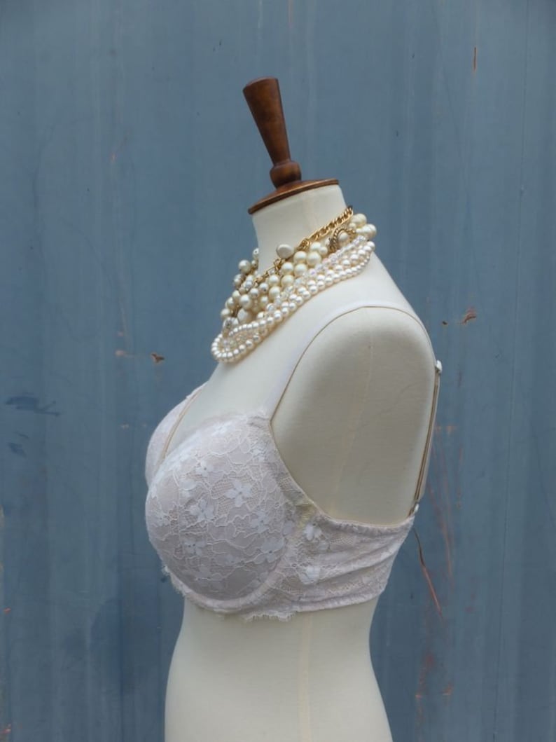 K&ouml;nnte beinhalten: Ein wei&szlig;er Spitzen-BH auf einer wei&szlig;en Schaufensterpuppe mit einer Perlen- und Goldkette. Die Puppe steht vor einem blauen Hintergrund.