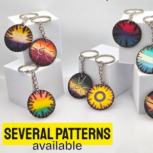 Peut inclure: Une collection de porte-clés circulaires présentant divers motifs peints à la main dans des couleurs vives, notamment des arcs-en-ciel, des soleils et des motifs abstraits. Chaque porte-clés a un anneau et une chaîne en métal. Texte : PLUSIEURS MODÈLES disponibles.