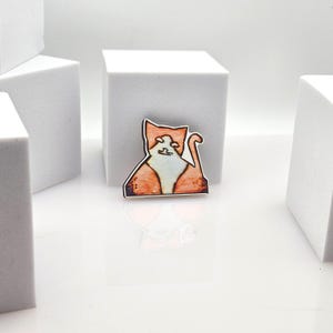 May include: A flat, decorative pin shaped like a sitting cat with orange and white fur. The cat has a distinct orange face, a white chest, and an orange body with a curved tail. The design features bold black outlines and a smooth, matte finish.