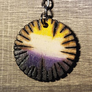 Puede incluir: Un colgante de llavero de madera con un diseño de la bandera del orgullo no binario. El colgante circular presenta un degradado de amarillo, blanco, morado y negro, con líneas grabadas alrededor del borde. Se adjunta un llavero de color plateado.