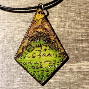 Peut inclure: Pendentif en bois en forme de losange, avec un motif vert et marron et des symboles gravés. Le pendentif est suspendu à un cordon noir. La partie supérieure est marron avec un motif de montagne, et le bas est vert avec des symboles gravés.
