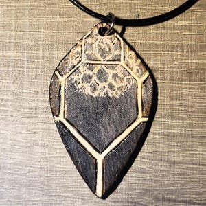 May include: A wooden pendant necklace with a geometric design. The pendant is a dark gray color with a lighter wood-colored geometric pattern. The pendant hangs from a black cord.