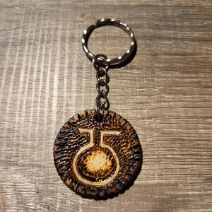Op de afbeelding: Een bruine leren sleutelhanger met een zilveren ketting en een ronde hanger met een wit symbool. Het symbool is een cirkel met een lijn erdoorheen en een kleinere cirkel bovenaan.