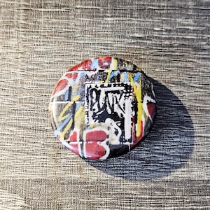 May include: A round, colorful button with a graffiti-style design. The button features a mix of red, blue, yellow, and black paint splatters and a central black and white graphic with text. The button is set against a textured, gray background.