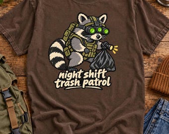 May include: A brown short-sleeve t-shirt featuring a graphic of a raccoon wearing tactical gear and night-vision goggles while holding a flashlight and a black trash bag. The text below the raccoon reads night shift trash patrol.