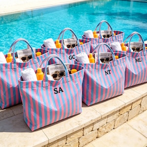May include: A collection of striped tote bags in pink and blue, each with a black initial. The bags are filled with white towels, sunglasses, sunscreen, and magazines. The bags are sitting by a pool.