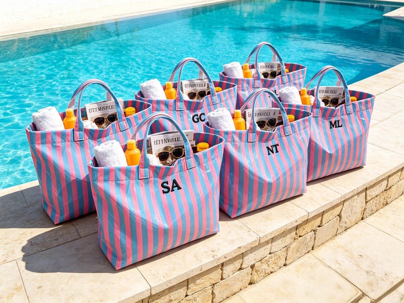 May include: A collection of striped tote bags in pink and blue, each with a black initial. The bags are filled with white towels, sunglasses, sunscreen, and magazines. The bags are sitting by a pool.