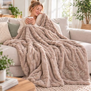 May include: A plush, textured beige faux fur throw blanket featuring an embroidered design of two interlocking hearts above the text "Mason & Lily" and "EST. 2023" in a cursive script, draped over a sofa to provide warmth and comfort.