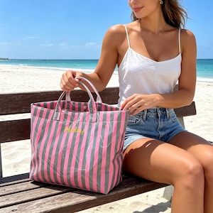 May include: A pink and gray striped tote bag with the name "Holly" embroidered in yellow. The bag has white handles and is sitting on a wooden bench. The background shows a beach and ocean.