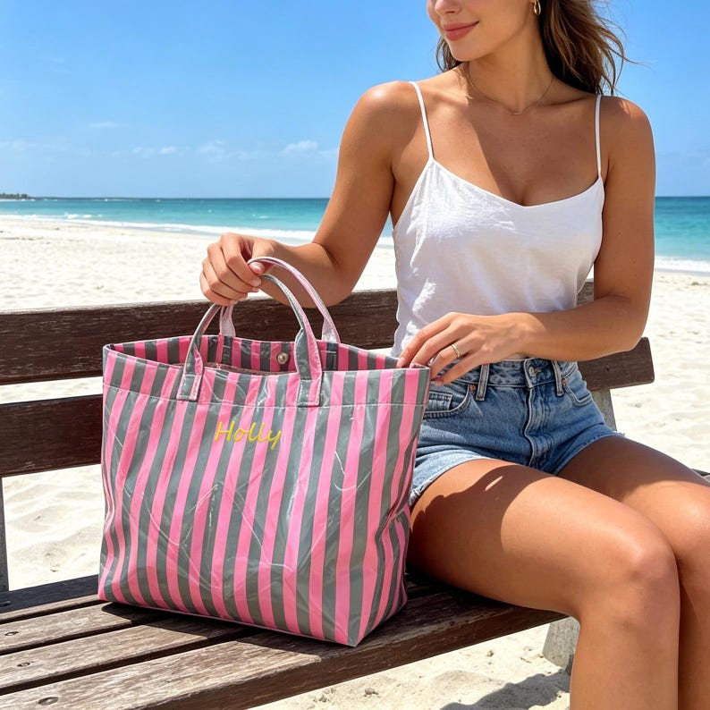 May include: A pink and gray striped tote bag with the name "Holly" embroidered in yellow. The bag has white handles and is sitting on a wooden bench. The background shows a beach and ocean.