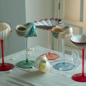Op de afbeelding: Een verzameling unieke schelpenglazen met gekleurde stelen en voetjes. De schelpen hebben verschillende vormen en kleuren, waaronder wit, turquoise en bruin. De stelen zijn rood, roze en groen. Verspreide parels zorgen voor een decoratief tintje.