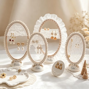May include: Cream-coloured oval earring displays with lace and pearl detailing, showcasing various earrings. The displays are labelled with names such as "Brielle", "Nikki", "Molly", and "Sara". A white ceramic dish and ring cones are also visible, all on a white surface.