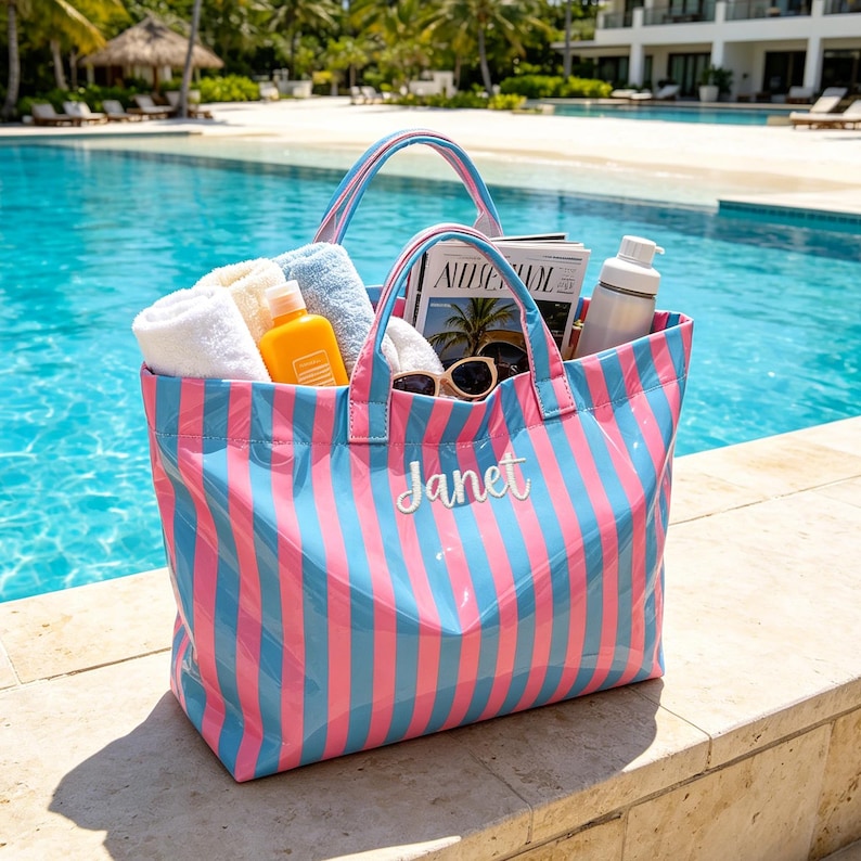 May include: A large, striped tote bag in pink and blue, filled with beach essentials like towels, sunscreen, sunglasses, and a magazine. The bag has a white embroidered name "Janet" on the front and is sitting by a pool.