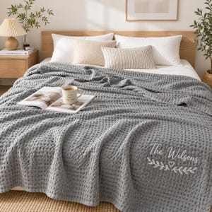 May include: A grey waffle-weave blanket draped over a bed, featuring the embroidered white text 'The Wilsons' above a decorative leaf and heart design. The blanket has a textured grid pattern and covers the bedding, which includes white pillows.