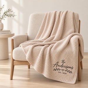 May include: A beige waffle-knit throw blanket featuring black printed text that reads The Andersons, EST. 2026, accompanied by a small heart icon and decorative leafy branch illustrations centred below the family name on the fabric.