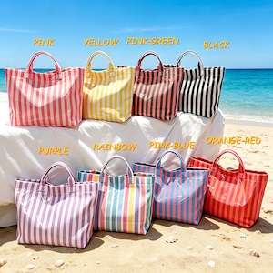 May include: A collection of striped tote bags in various colors, including pink, yellow, pink-green, black, purple, rainbow, pink-blue, and orange-red. Each bag has a rectangular shape with a matching handle. The bags are displayed on a sandy beach with the ocean in the background.