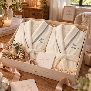 May include: Two cream-colored plush robes with grey piping, embroidered with Mr. Taylor and Mrs. Taylor, presented in a gift box with a dried floral bouquet, a card reading With Love, and the text A MORE BEAUTIFUL LIFE TOGETHER on the box edge.