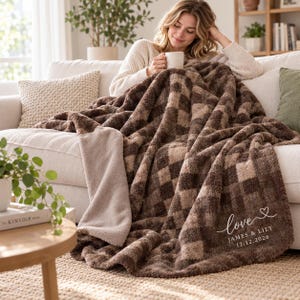 May include: A plush, textured throw blanket featuring a brown and beige checkered pattern. The blanket includes white embroidered text that reads love JAMES & LILY 12.12.2026 with a small heart icon. The reverse side is a solid light beige colour.