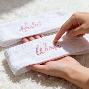 Peut inclure: Deux bandeaux blancs en éponge avec des noms brodés en rose. L'un dit "Harleen" et l'autre "Winda". Les bandeaux sont tenus par les mains d'une personne. Fond blanc et moelleux.