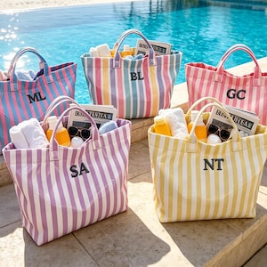 May include: Five striped tote bags in various colors, including pink, blue, and yellow, are filled with beach essentials. Each bag has a different initial: ML, BL, GC, SA, and NT. The bags are sitting by a pool.