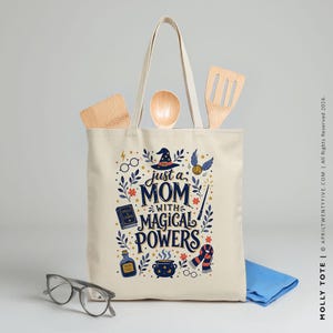 Może przedstawiać: Kremowa torba z napisem "just a MOM with MAGICAL POWERS" w kolorze granatowym, czerwonym i złotym. Torba posiada kapelusz czarownicy, różdżkę i inne magiczne elementy. Drewniane przybory kuchenne znajdują się wewnątrz torby, a przed nią para okularów i niebieska szmatka.