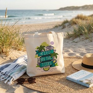 Può includere: Una borsa tote in tela beige con la scritta "#TIED BY THE TIDES" in verde acqua e verde, con palme e una scena di spiaggia. La borsa è su una spiaggia sabbiosa con un cappello di paglia, occhiali da sole e un libro intitolato "Coastal Living".