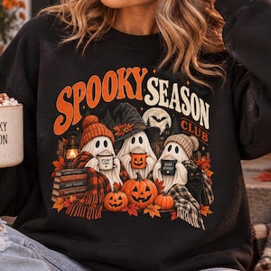 Puede incluir: Una sudadera negra de cuello redondo con un estampado gráfico con el texto "SPOOKY SEASON CLUB" sobre tres fantasmas con sombreros, sosteniendo tazas y sentados con calabazas, libros y un gato negro. Se sostiene una taza con el texto "SPOOKY SEASON".