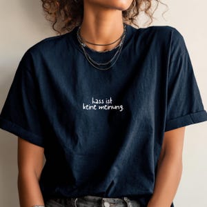 May include: Navy blue t-shirt with the German phrase "hass ist keine meinung" in white script. The shirt has short sleeves and a relaxed fit. The model is wearing multiple layered necklaces.