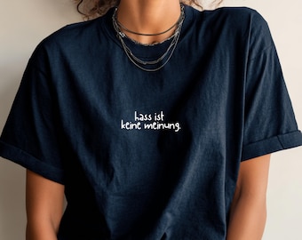 May include: Navy blue t-shirt with the German phrase "hass ist keine meinung" in white script. The shirt has short sleeves and a relaxed fit. The model is wearing multiple layered necklaces.