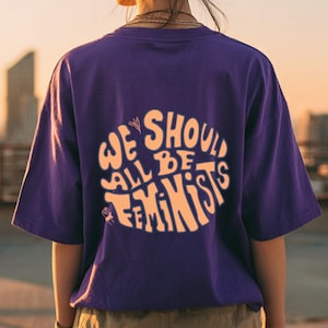 Könnte beinhalten: Ein lila T-Shirt mit dem Text "WE SHOULD ALL BE FEMINISTS" in einem kreisförmigen Design. Der Text ist hellorange. Das Shirt hat kurze Ärmel.