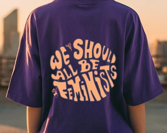 May include: A purple t-shirt with the text "WE SHOULD ALL BE FEMINISTS" in a circular design. The text is in a light orange color. The shirt has short sleeves.