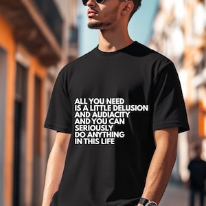 Könnte beinhalten: Schwarzes T-Shirt mit weißem Text: "ALL YOU NEED IS A LITTLE DELUSION AND AUDACITY AND YOU CAN SERIOUSLY DO ANYTHING IN THIS LIFE." Das Shirt hat einen klassischen Rundhalsausschnitt.