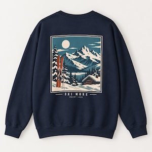 Op de afbeelding: Een marineblauw sweatshirt met ronde hals en een rechthoekige afbeelding op de achterkant. De illustratie toont een besneeuwd berglandschap met een hut, dennenbomen en een paar oranje ski's. De tekst luidt "SKI MORE" en "WORRY LESS".