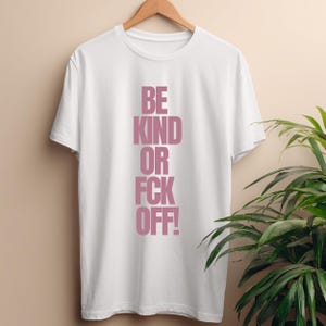 Könnte beinhalten: Weißes T-Shirt mit dem vertikalen Text "BE KIND OR FCK OFF!" in Rosa. Das Shirt hängt an einem Holzbügel vor einem neutralen Hintergrund, mit einer grünen Pflanze auf der rechten Seite.