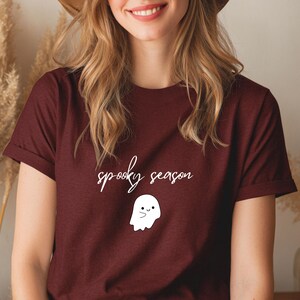 Könnte beinhalten: Burgunderfarbenes T-Shirt mit dem Schriftzug "spooky season" in Schreibschrift und einer weißen Geistergrafik. Der Geist hat ein lächelndes Gesicht. Das Shirt ist aus weichem Material.