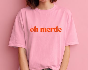 May include: A light pink t-shirt with the words "oh merde" in orange lettering. The shirt is a crew neck style and appears to be made of a soft material. The background is a solid pink color.